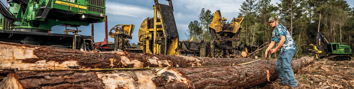 A logger measures a tree while Deere forestry equipment sits in the background.