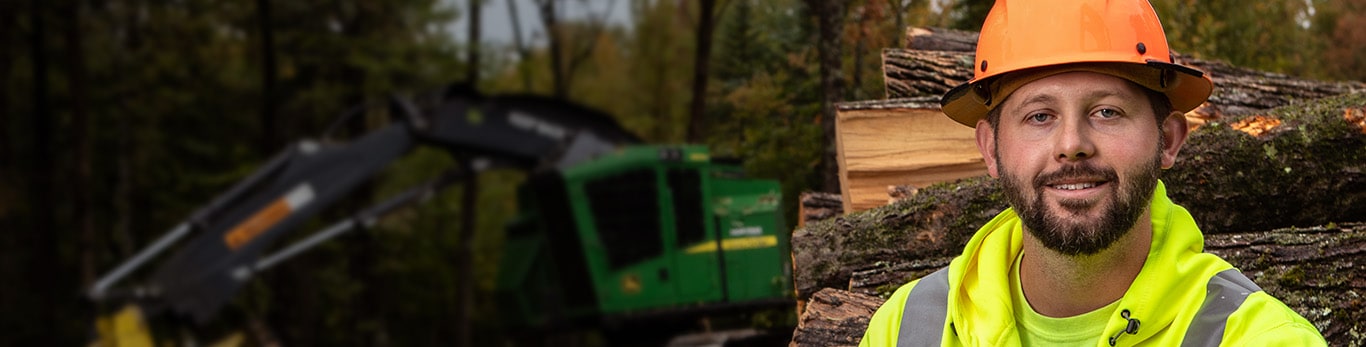 Zachary Emerson sitting on a stack of logs and in the background is a John Deere 853M Tracked Feller Buncher with a FR22B Felling Head.