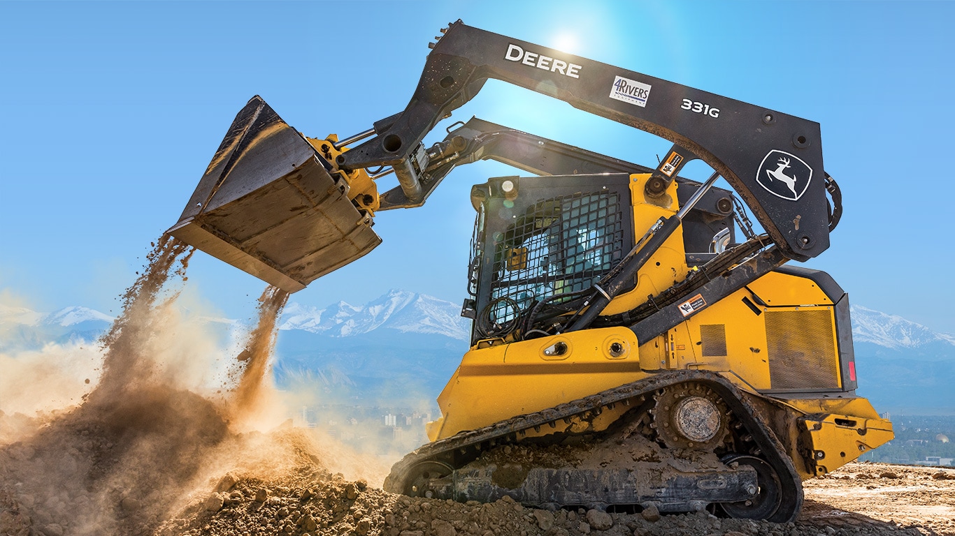 With the Rocky Mountains in the background a 331G Compact Track Loader dumps a bucketload of dirt at a jobsite.