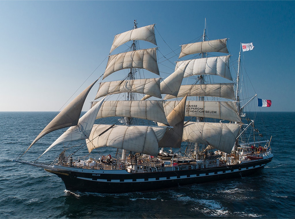 A large three-masted tall ship with white sails and a French flag flying from the mast sails on the ocean.