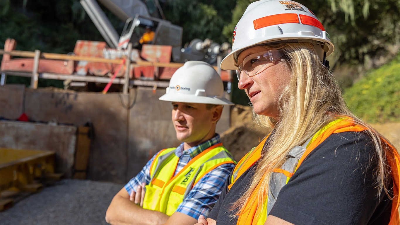 Two construction workers wearing safety vests, goggles, and hard hats standing in a construction worksite looking off into the distance