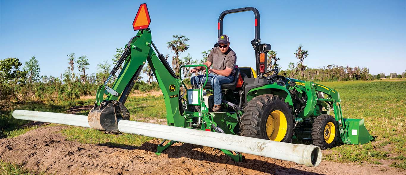 A green backhoe attached to a green compact tractor holds a large pipe above a trench.