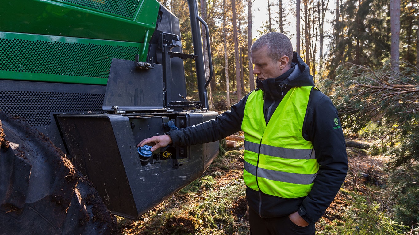 Technician servicing John Deere H-Series forestry machine with open engine compartment.