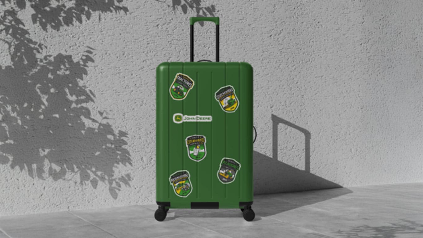 Fondo gris con una maleta verde decorada con pegatinas de los Desafíos John&nbsp;Deere.