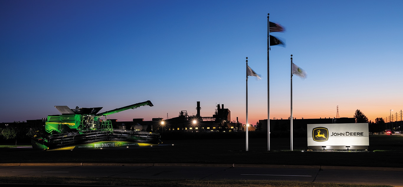 Imagen nocturna al atardecer de una instalación industrial de John Deere, con maquinaria exhibida, banderas en mástiles y el logotipo iluminado.