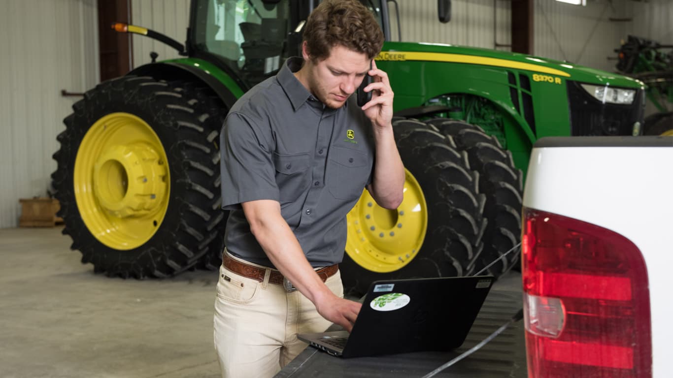 person working in laptop and talking on phone with John Deere tractor in background