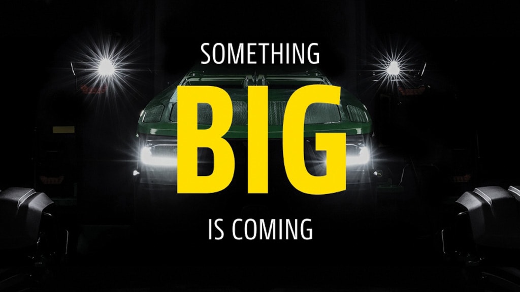 Dark promotional image of a machine with bright headlights emerging from shadows and text saying 'SOMETHING BIG IS COMING'