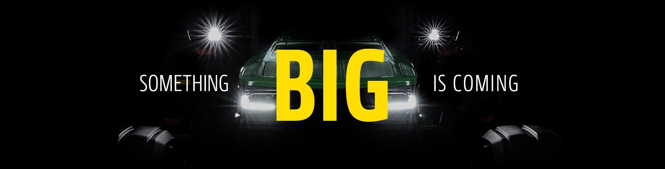 Dark promotional image of a machine with bright headlights emerging from shadows and the text 'SOMETHING BIG IS COMING'