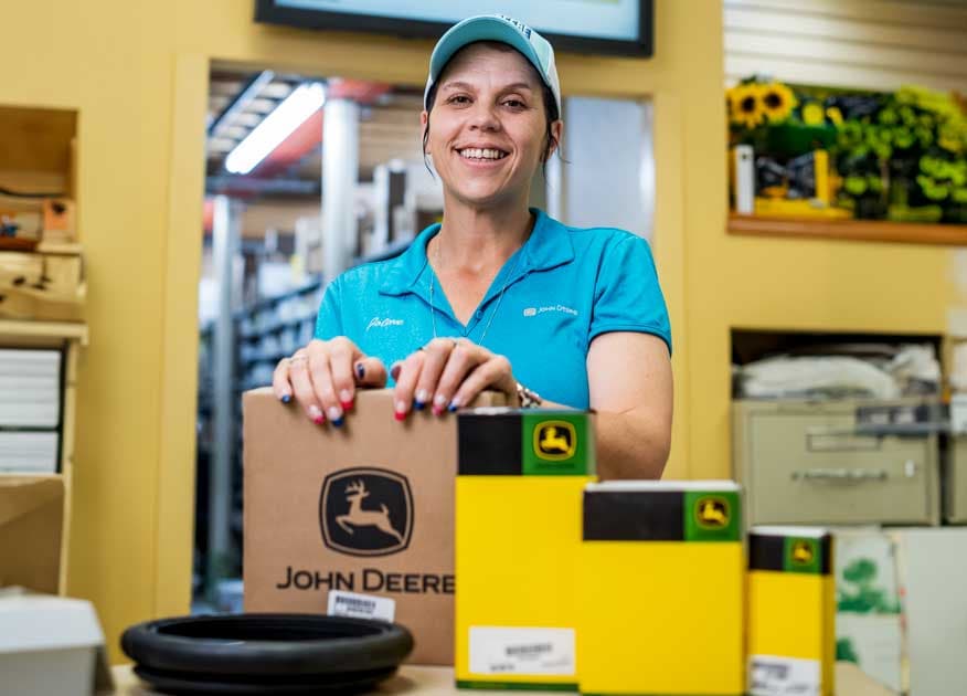 Woman in a light blue polo and ball cap behind a desk at a dealership with John Deere Parts in their boxes sit in front of her on the desk