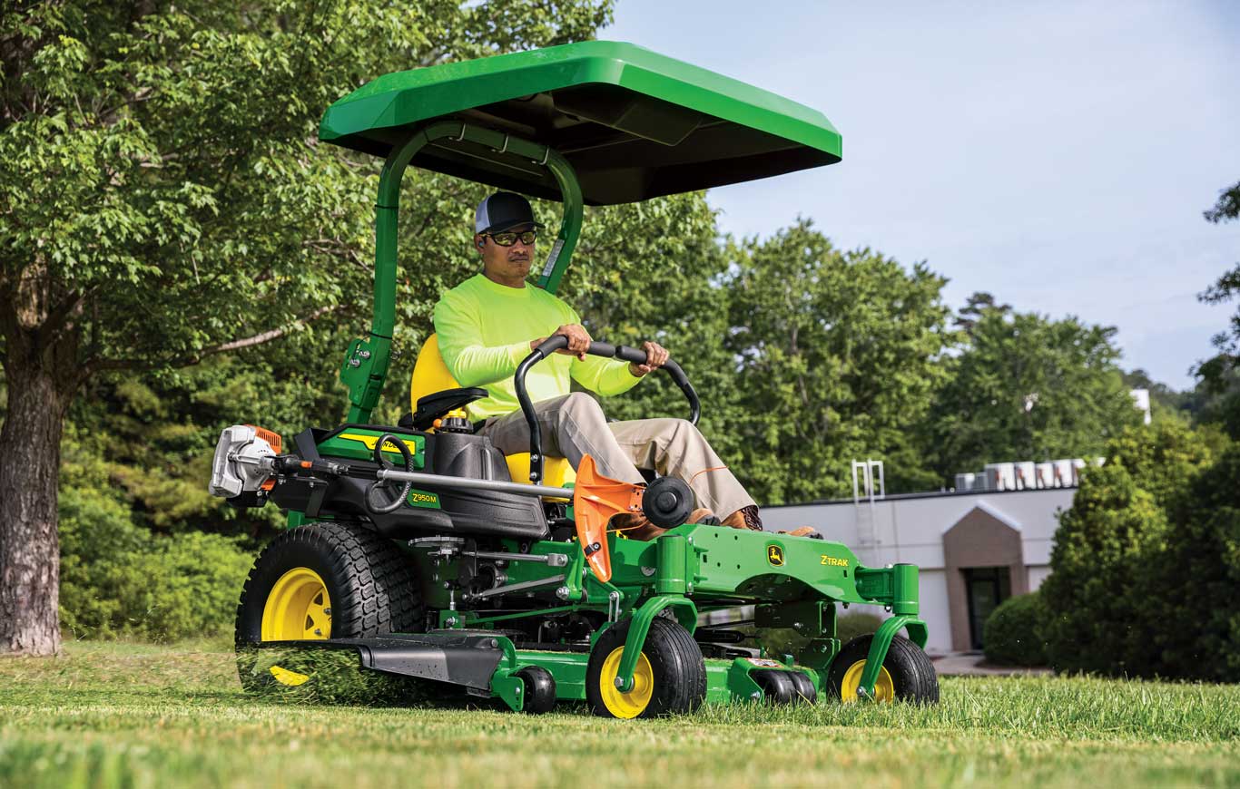 Man with neon shirt and safety sun glasses operating a John Deere Commercial Zero Turn Z950M ZTrak with 60 in deck, ShadePro canopy, and an attachmentment with a Stihl edger in front of a business