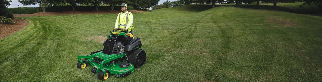Man with saftey glasses and a yellow safety vest operating a John Deere Commercial Stand-On Mowers Q850R QuikTrak with tweel tires in an open grass/park area (some contents of this images may be AI generated)