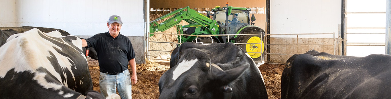 Farmer in barn with black and white cows; green tractor with yellow wheels in background.
