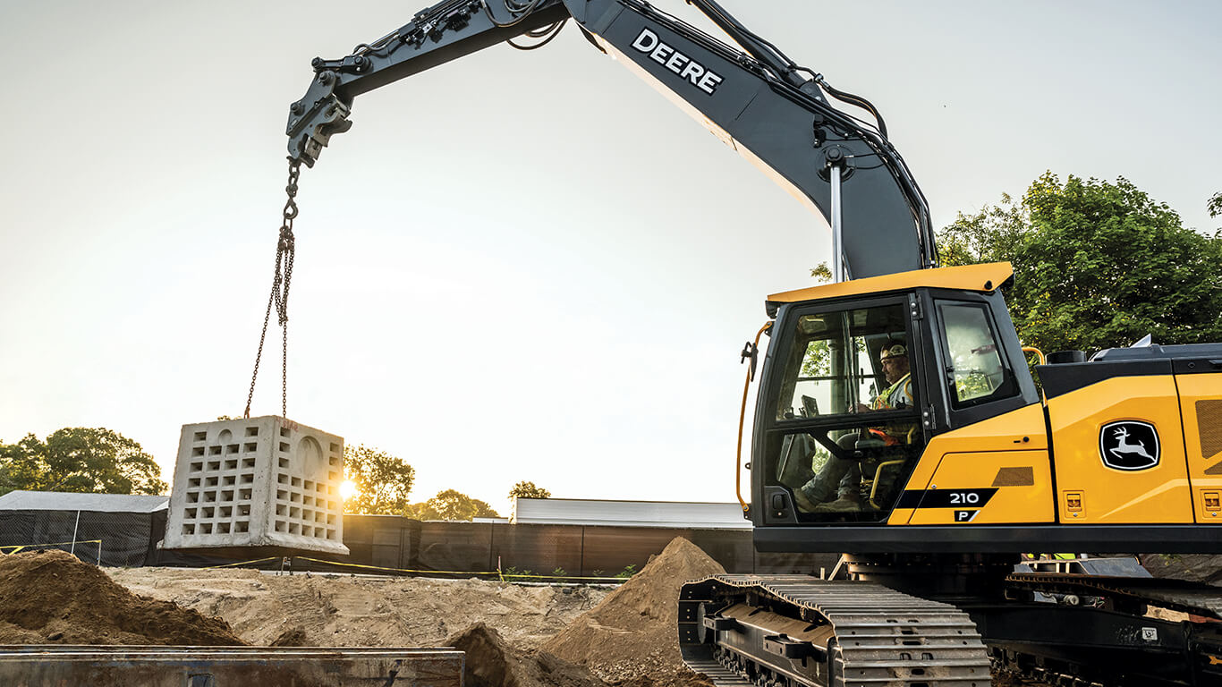 John&nbsp;Deere excavator lifting concrete blocks with a chain at a construction site, dirt piles in foreground.