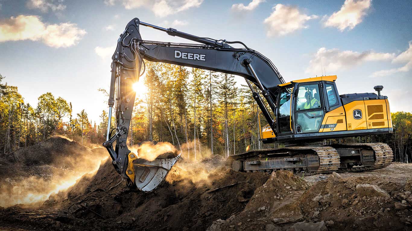 John&nbsp;Deere excavator digging soil at a construction site, sunlight through trees in the background.
