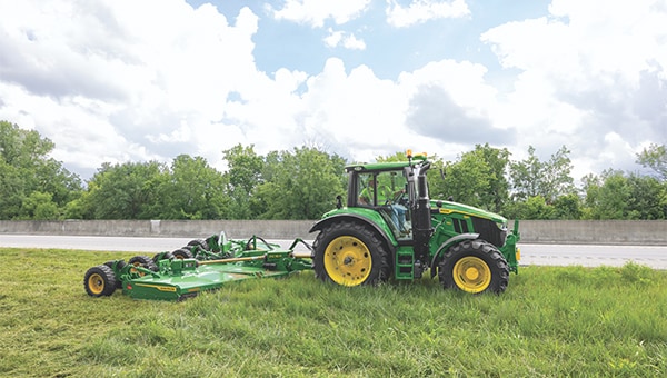 Green tractor pulling rotary mower cutting tall grass beside a roadway under cloudy sky.