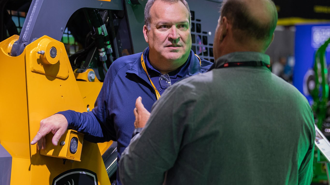 Two individuals talk indoors next to a yellow skid steer.