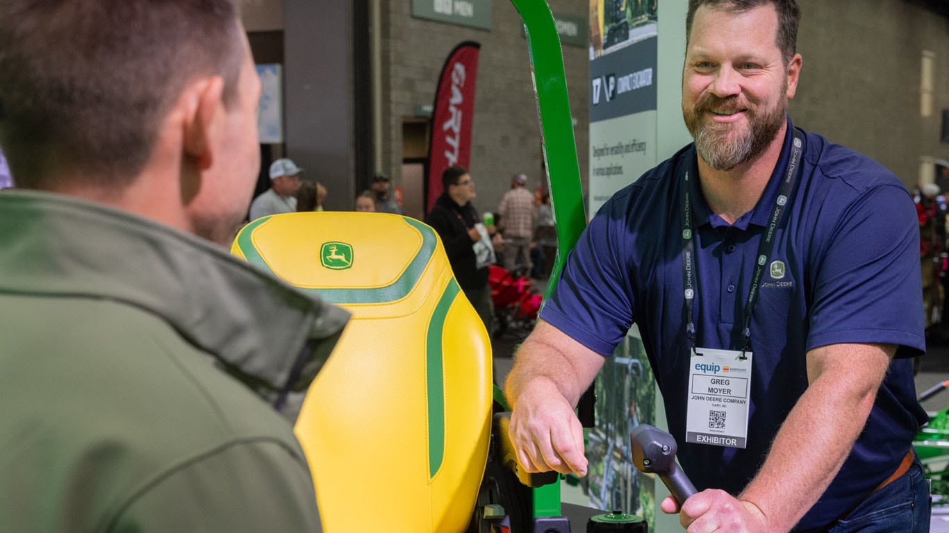 Indoors, an equip expo exhibitor talks to someone next to a green zero-turn mower.