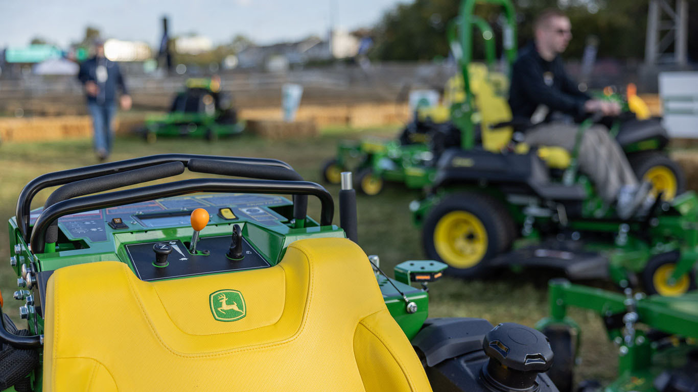 Close-up shot of a green standing mower control panel with various blurry Deere mowers in the background.