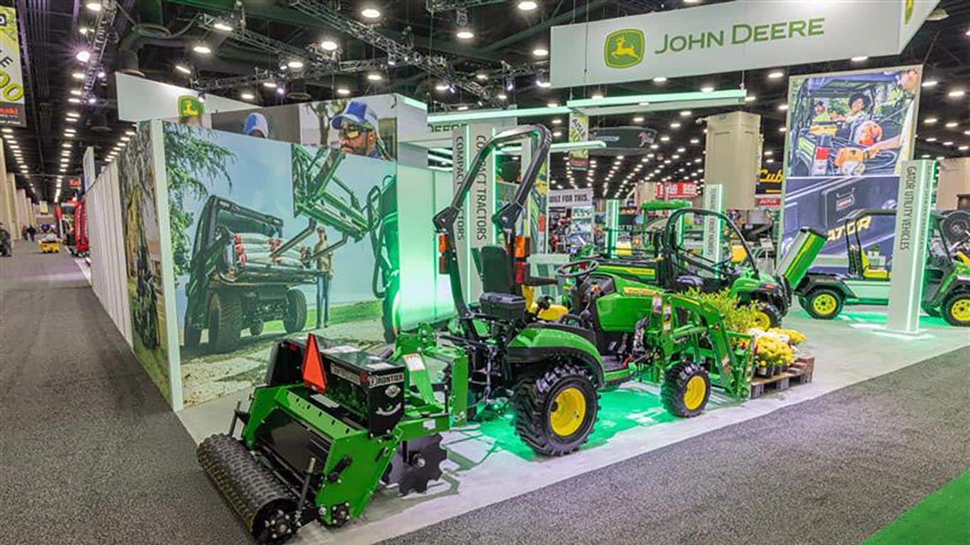 Part of the indoor Deere booth, a compact tractor has a pallet fork holding flowers and a seeder on the back.