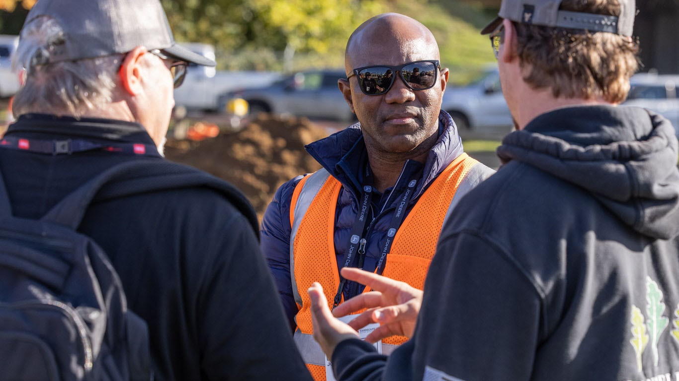 Outdoors, two individuals talk to someone in an orange work vest.