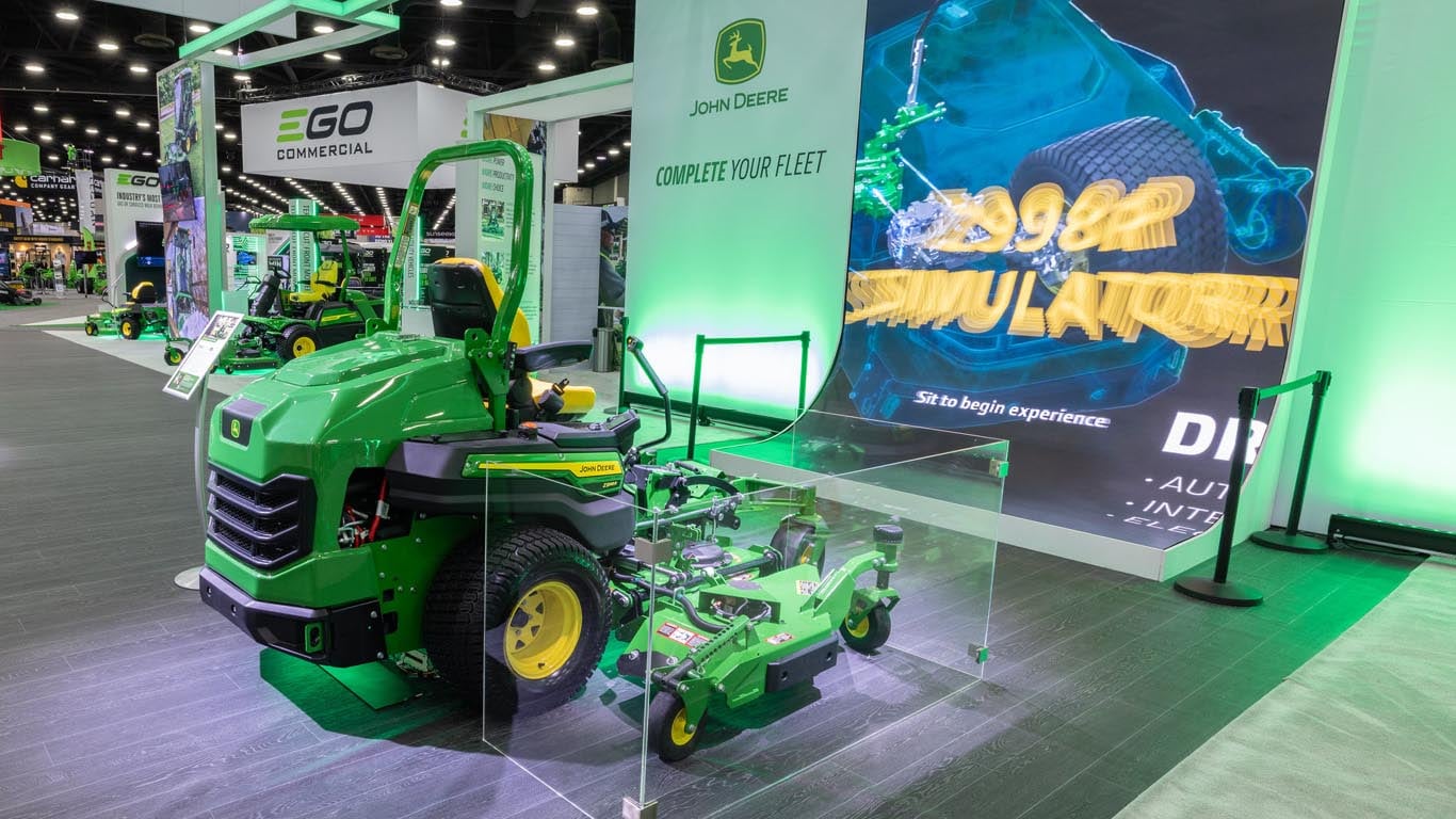 Indoors a green 100-in. zero-turn mower, the wingdecks boxed in with glass, sits in front of a tall video screen.