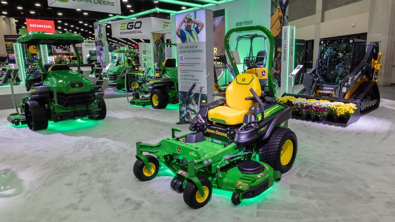 Indoors two zero-turn mowers rest of either side of a sign saying how to get a free Deere hat.