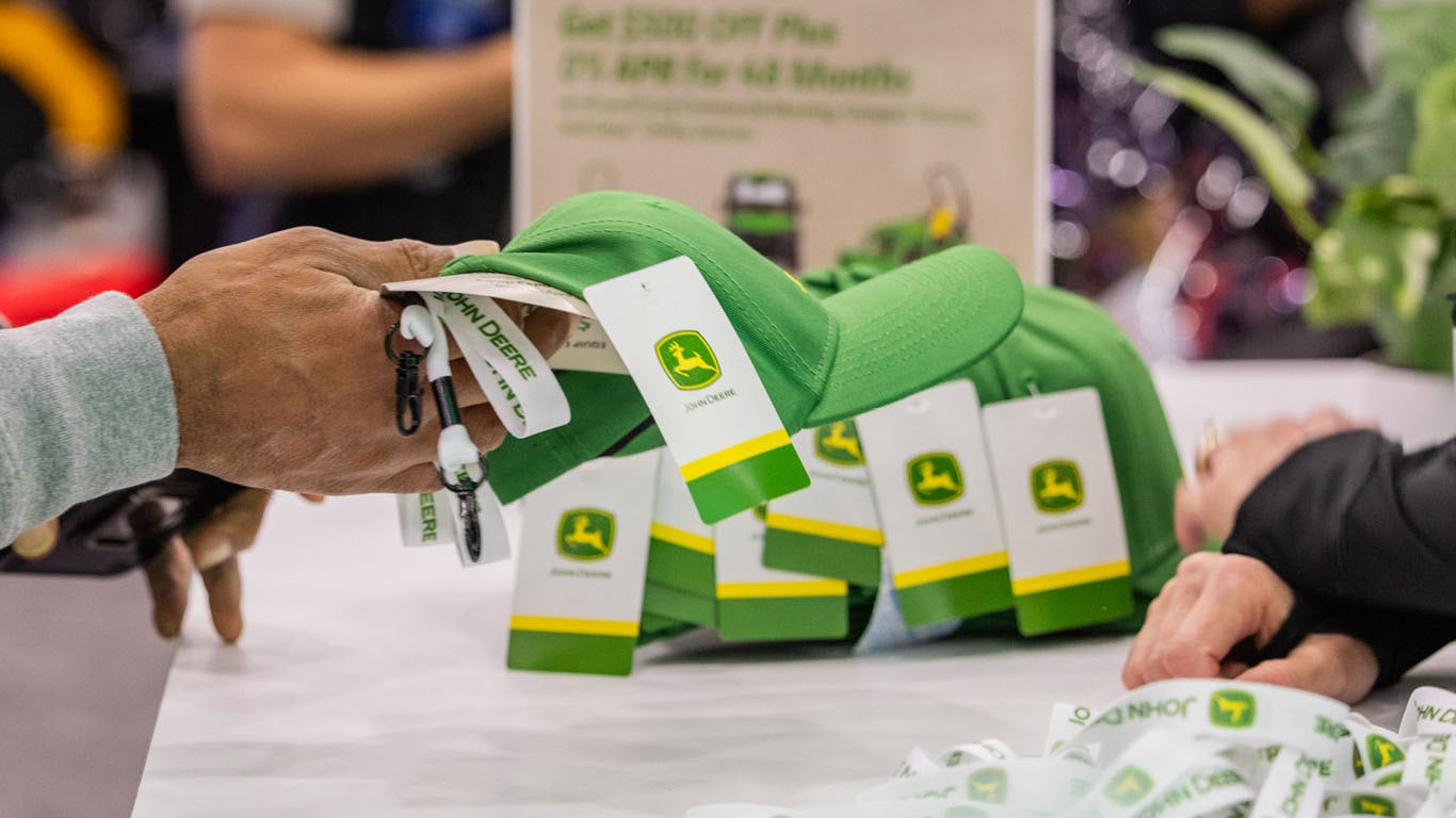 A hand picks up a green Deere hat from a line of them on a counter.