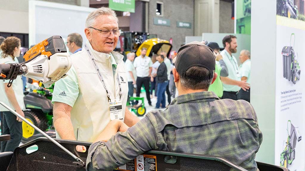 Two individuals talking at a booth while other expo attendees are walking around in the background.