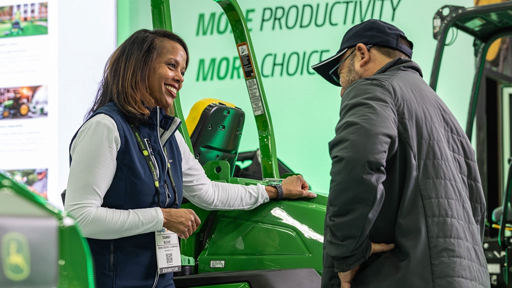 Two people talk next to a green zero-turn mower at an indoor booth.