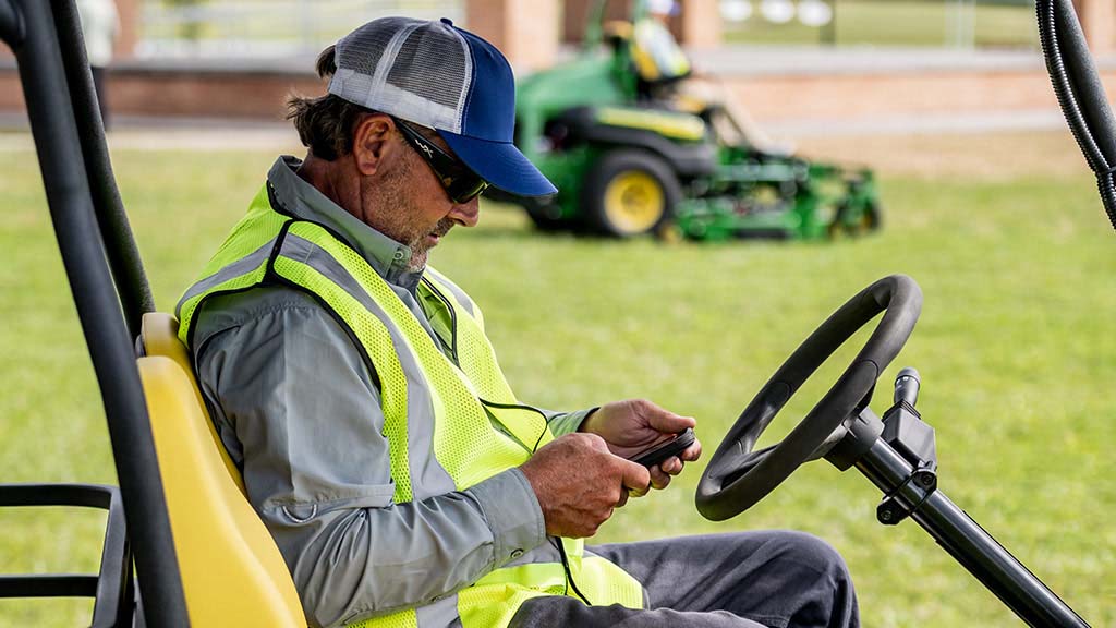 A person in a work vest sitting on a yellow gator seat while looking down at their phone.