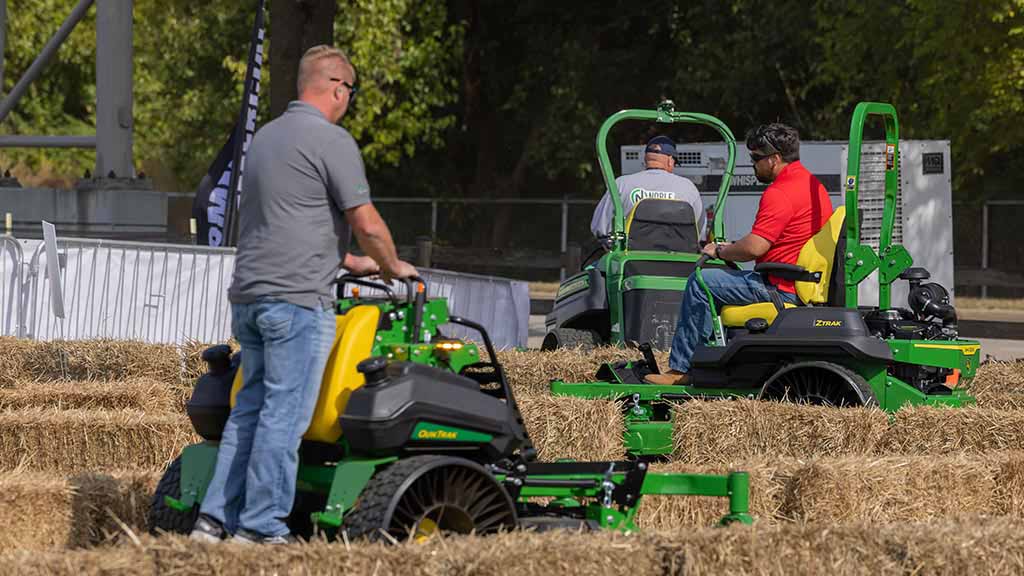 Three individuals riding three different green mowers around an outdoor track lined with hay bales.