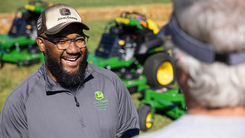Two people talk outside with green stand-on mowers in the background behind them.