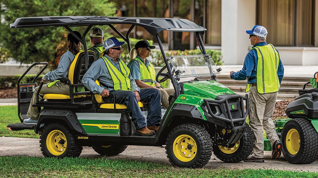 Four campus workers in safety vests in a green electric Gator parked as a fifth worker walks by.