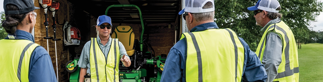Workers in safety vests stand at the open back of a trailer with lawn maintenance equipment while one person demonstrates a zero-turn mower.