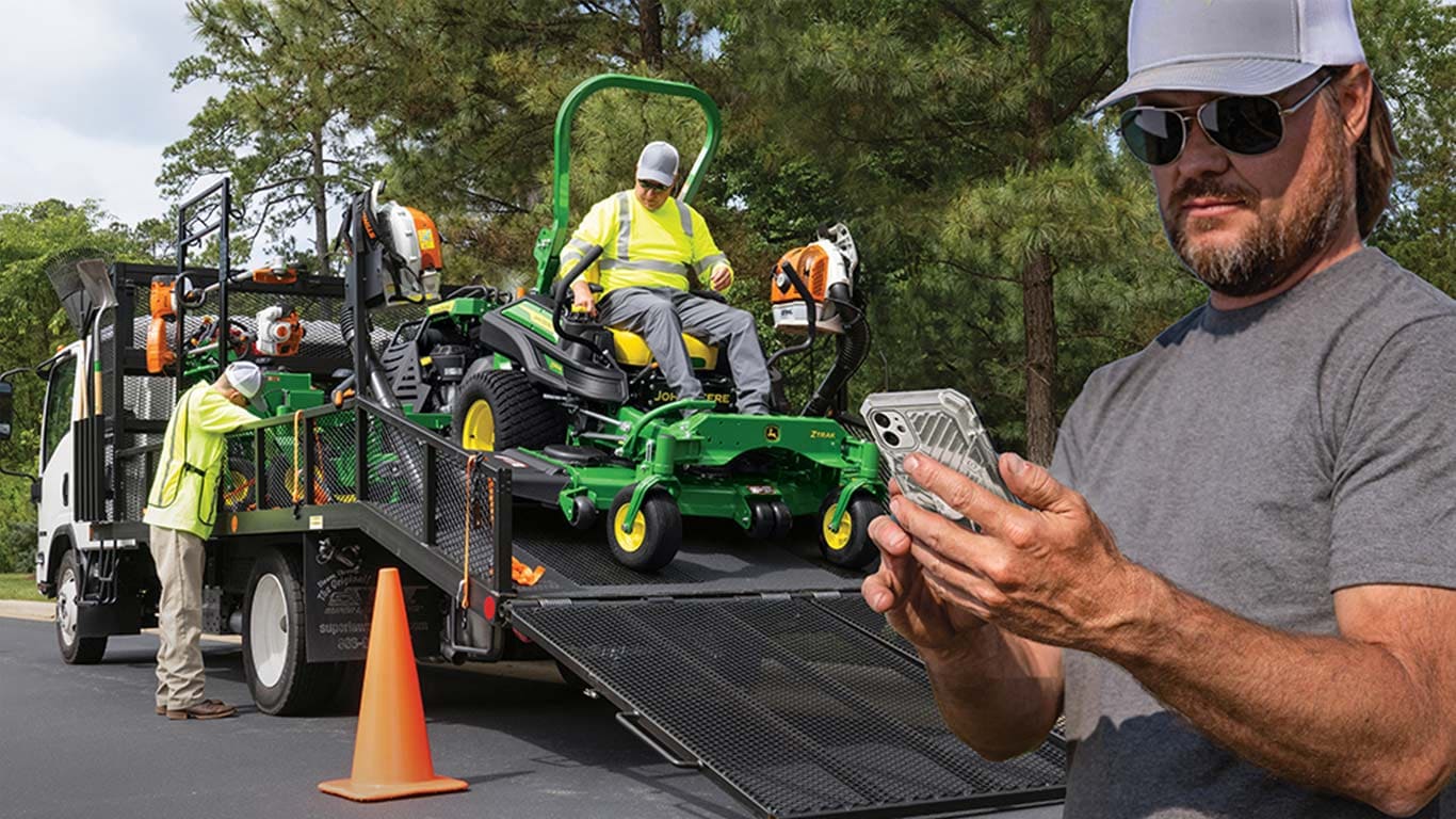 Man looking at his smartphone while landscape professionals take John Deere equipment off of a flatbed truck in the background.