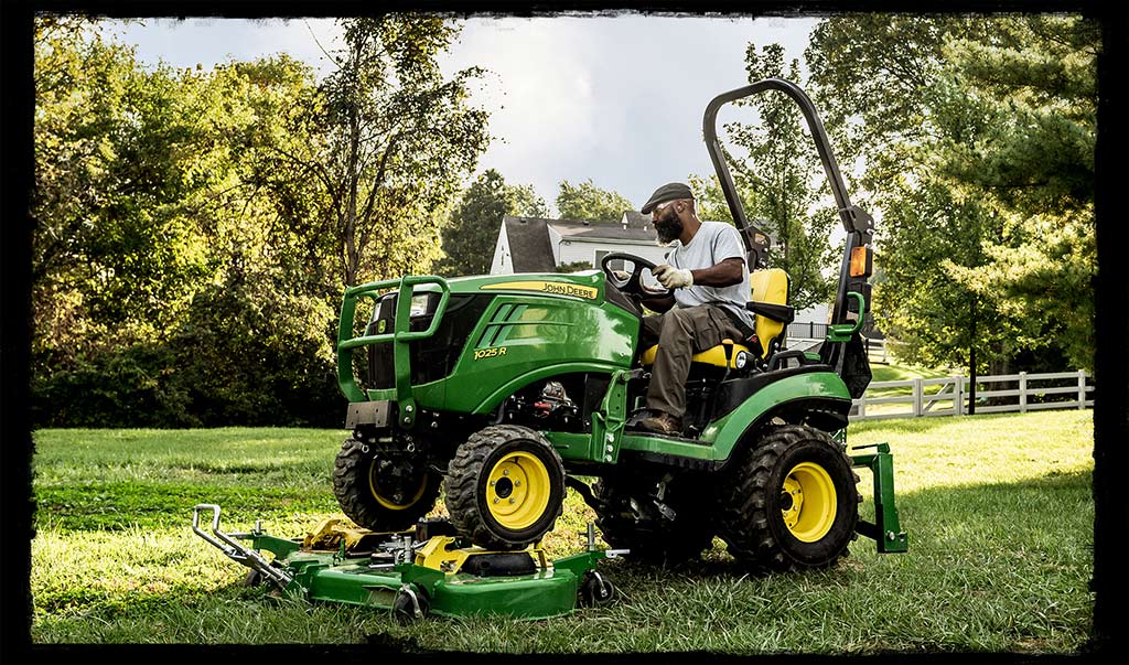 Man on yellow and green John Deere 1 Series tractor is in a green grass yard with a fence and large trees in the background. 