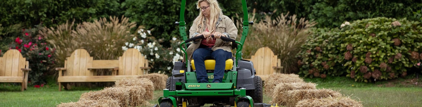 Woman with blonde hair and a gray shirt and jeans rides a green and yellow John&nbsp;Deere riding lawn mower cutting the grass. There is a wooden bench in the background and hay bales on either side of the mower. 