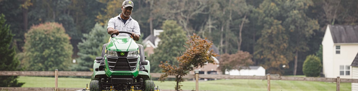 A man with a gray beard wearing a light gray shirt and a black baseball cap rides on a green and yellow John Deere lawn mower in a grass yard with a wooden fence in the background. 