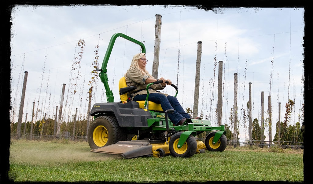 Woman with blonde hair and a gray shirt and blue jeans rides a green and yellow John Deere riding lawn mower cutting the grass. 