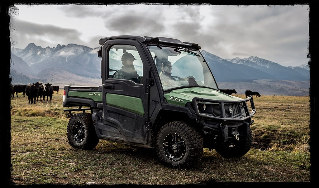A black and dark green John Deere utility vehicle rides though the grass with two people’s figures showing inside the cab. Snowcapped mountains are in the background and cows.
