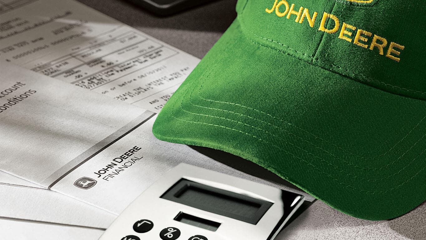 Green John Deere hat resting on a table next to printed financing documents and calculator.