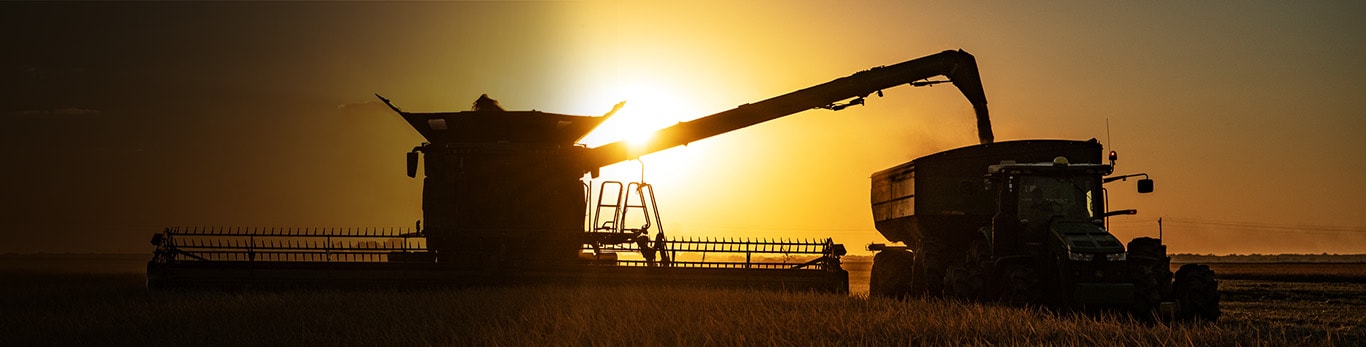Silhouetted John Deere combine unloading into a grain cart at sunset in a golden field.