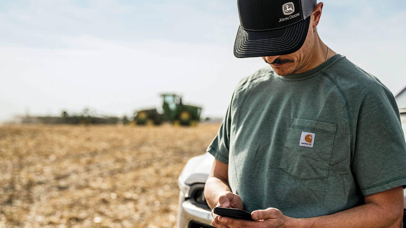 Person looking down at his phone in a field with a blurry green tractor in the background.
