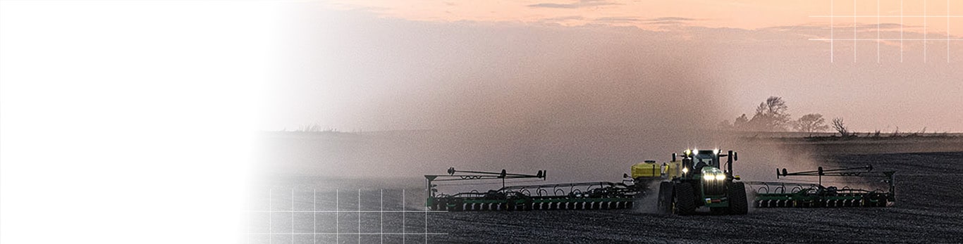 A tractor pulling a wide planter across a field at dusk, creating a trail of dust.