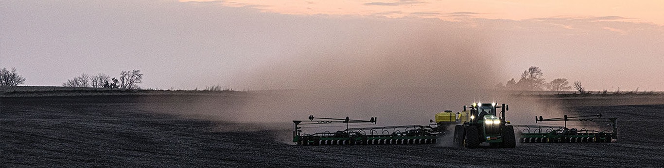 A tractor pulling a wide planter across a field at dusk, creating a trail of dust.