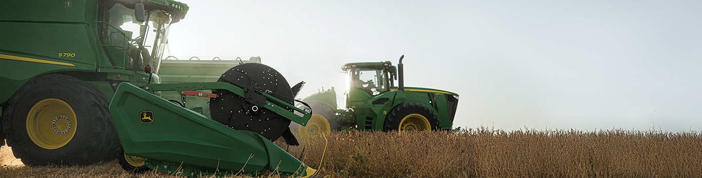 A John Deere combine unloading grain beside a John Deere tractor and grain cart in a soybean field.