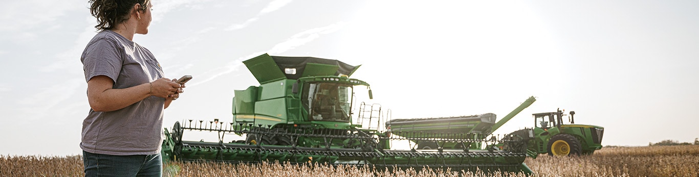 A person standing in a field while a John Deere combine and tractor operate in the background.