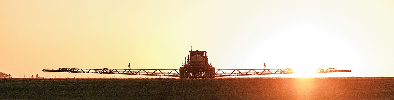 A sprayer with its boom extended across the field at sunrise.
