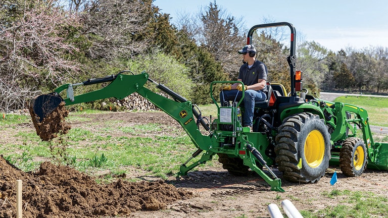 A person with ear and eye protection using a green backhoe to dump dirt in a pile