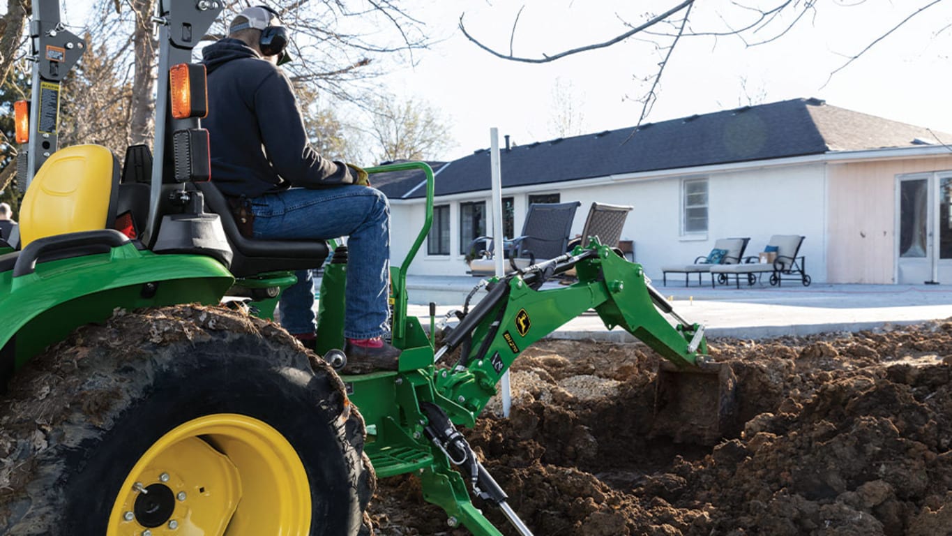 A person with ear and eye protection using a green backhoe to dump dirt in a pile